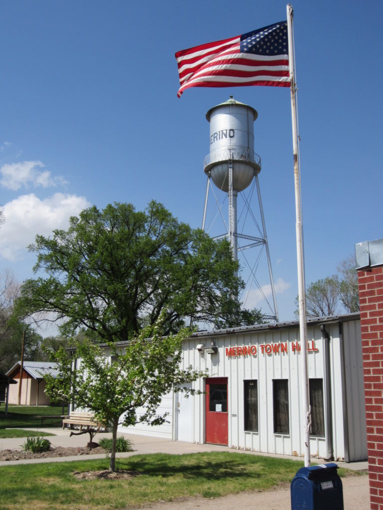 Image of Flag flying in front of the Merino town hall with the old water tower in the background.